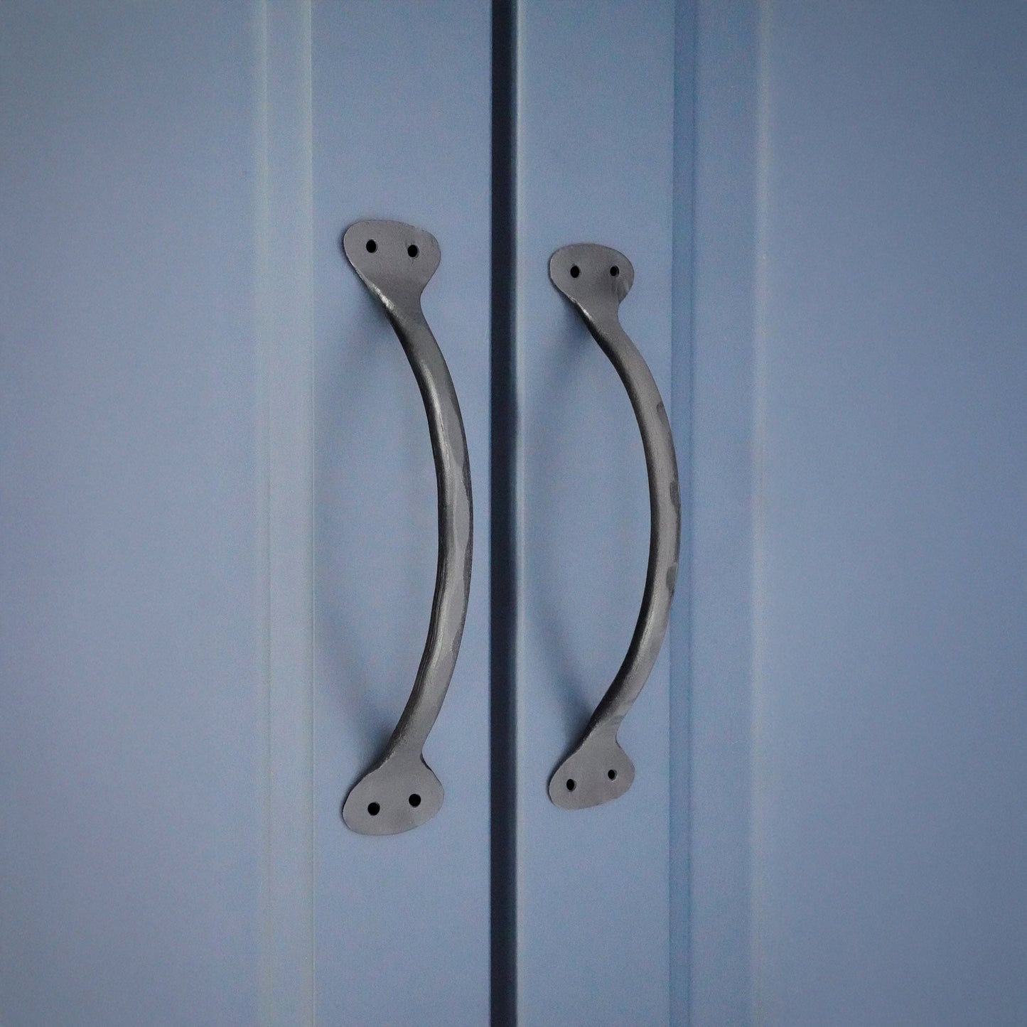 Spira Brass Hammered Pull Handle in a metallic finish, featuring a curved design with two mounting holes on each end, shown applied to a blue cabinet door.