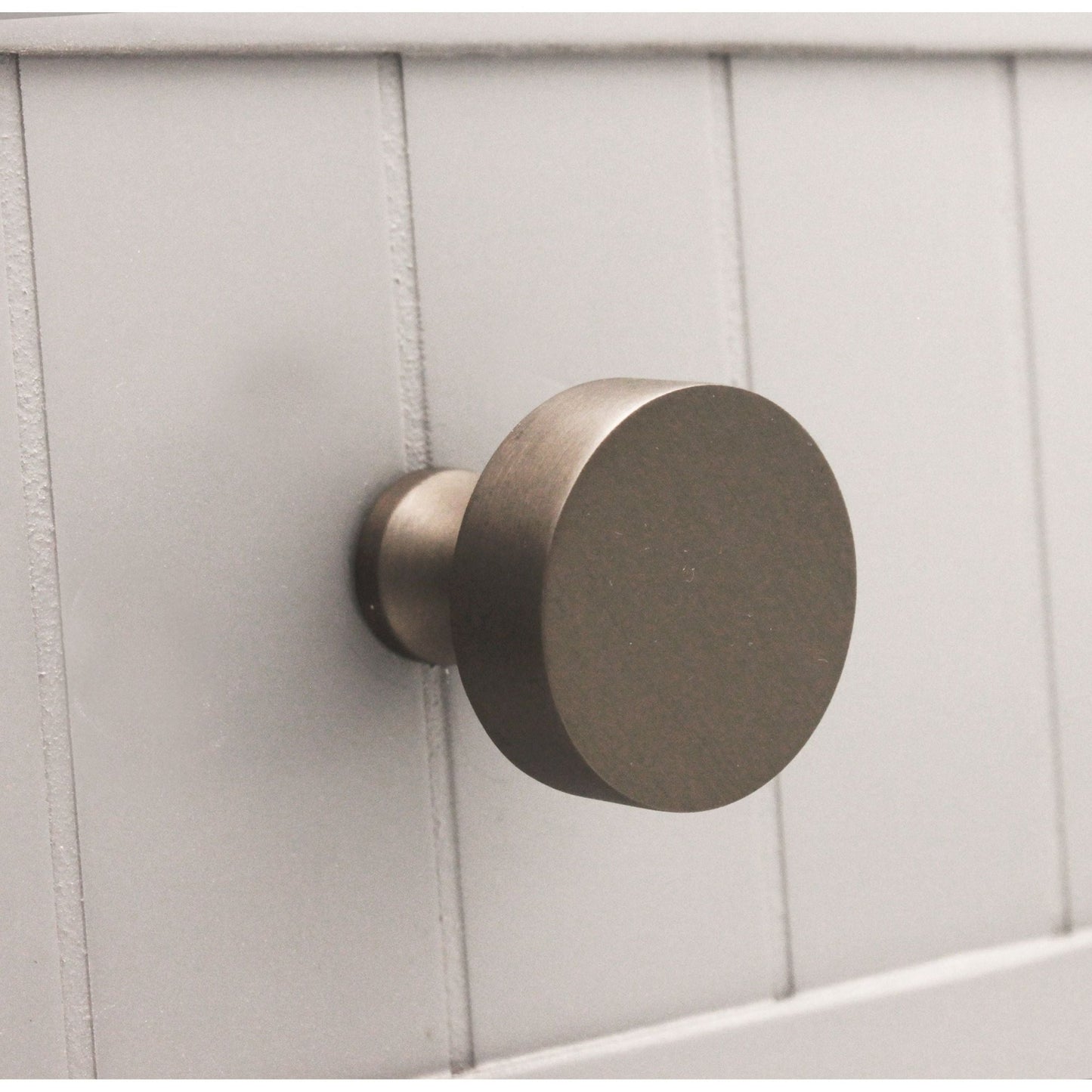 Spira Brass Round Cupboard Knob in a dark bronze finish, featuring a simple circular design and mounted on a wooden panel.