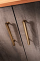 Alexander & Wilks Vesper Hex T-Bar Cabinet Pull in a brushed gold finish, featuring a sleek, elongated design mounted on two cabinet doors made of textured gray wood.