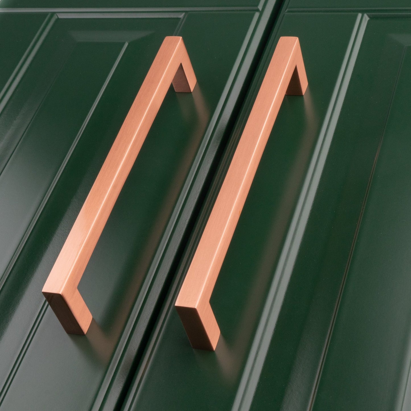 Carlisle Brass Block Handle in copper finish, featuring a rectangular design, mounted on a green cabinet door. The pulls are positioned side by side, showcasing their straight lines and geometric shape.