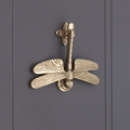 Spira Brass Dragonfly Door Knocker in a brass finish featuring detailed dragonfly wings and body with a mounted design. The piece is installed on a panel surface, showcasing its intricate textures.